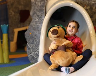 Ein Mädchen sitzt auf einer Rutsche mit einem Kuscheltier im Spielzimmer vom Mondi Resort Oberstaufen.