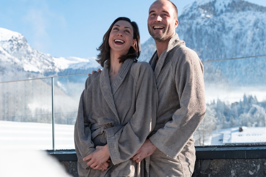 Hotel Franks Winter Ein Paar im Bademantel vor schneebedeckten Bergen im Hotel Franks in Oberstdorf.