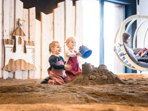 Zwei kleine Kinder spielen im Indoor-Sandkasten im Allgäuer Berghof in Gunzresried während eine Frau dahinter in einem Hängesessel liest