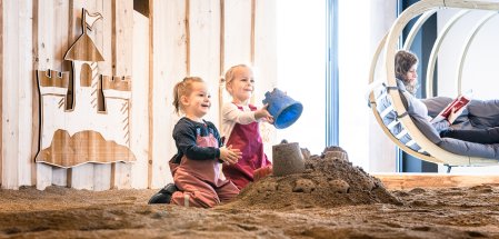 Allgäuer Berghof Sandkasten Zwei kleine Kinder spielen im Indoor-Sandkasten im Allgäuer Berghof in Gunzresried während eine Frau dahinter in einem Hängesessel liest