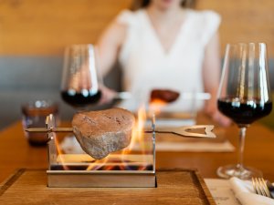 Ein Stück Fleisch grillt am Liquid Grill auf dem Tisch eines Gastes und wird von zwei Weingläsern flankiert