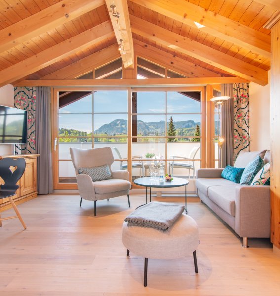 Einblick in eines der Zimmer im Hotel Hahnenköpfle in Oberstdorf mit Blick auf Sitzgelegenheiten und die breiten bodenlangen Fenstern mit dem tollen Ausblick auf die Allgäuer Berge