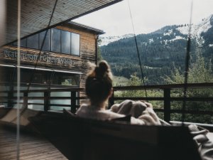 Eine Frau liegt auf einer Liege im Außenbereich des Wellnessbereichs im Naturhotel Chesa Valisa in Hirschegg und schaut auf die noch etwas verschneiten Berge