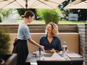 Eine Mitarbeiterin bringt einer Frau auf der Terrasse im Rosenalp Gesundheitsresort & Spa in Oberstaufen eine Speise