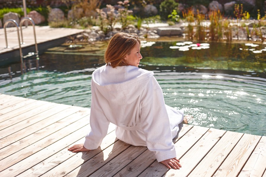 Eine Frau im Bademantel sitzt am Rande des Naturpools im Außenbereich des Biohotel Eggensberger in Hopfen am See / Füssen und kühlt ihre Beine