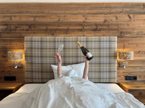 Eine Person liegt im Bett in einem Zimmer des Biohotel Eggensberger in Hopfen am See und hält eine Sektflasche und ein Glas in die Höhe.