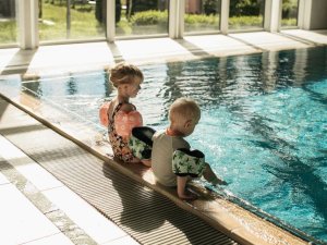 Zwei Kinder mit Schwimmflügeln sitzen am Beckenrand des Hallenbads des Mondi Resort in Oberstaufen.
