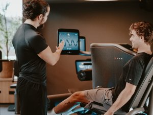 Ein Fitnesstrainer des Bergkristall - Mein Resort im Allgäu in Oberstaufen weist jemanden an den Geräten des Fitnessraums im Hotel ein.