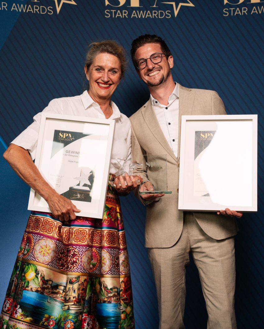 Hotel Franks Auszeichnung Zwei Personen vom Hotel Franks stehen vor einer offiziellen Award-Leinwand und halten die Preise sowie Auszeichnungen in der Hand
