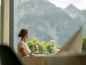 Eine Frau sitzt am Fenster im Restaurant des Hotel Prinz-Luitpold-Bads in Bad Hindelang und hält ein Glas Sekt währen sie aus den bodentiefen Fenstern schaut