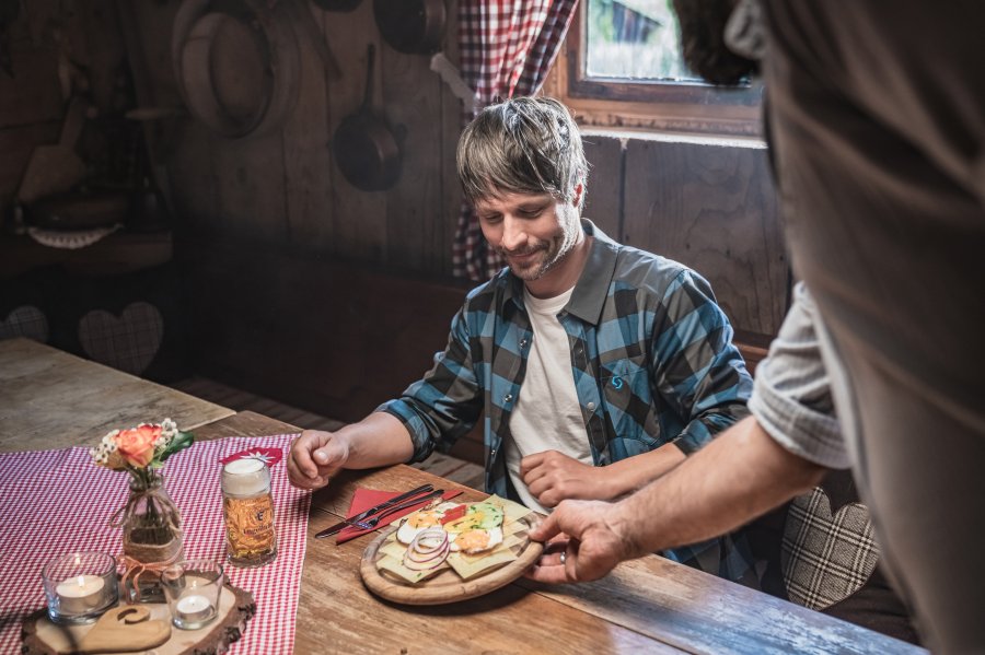 Allgäu GmbH Hütte Ein Mann erhält in einer urigen Hütte ein Brotzeitbrett mit Leckerrein serviert