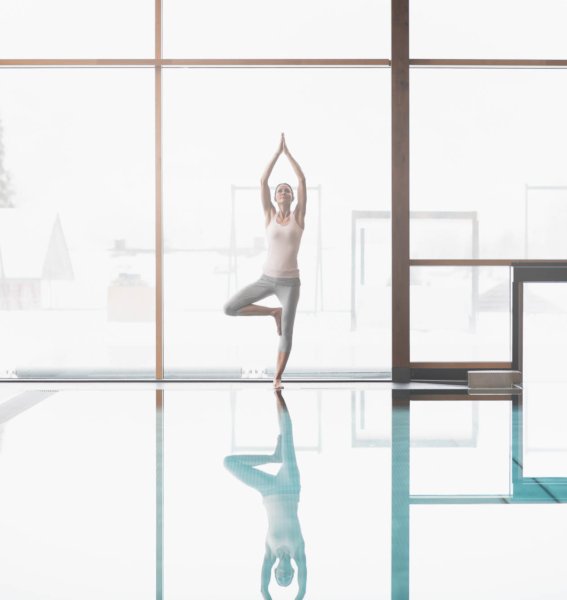 Eine Frau führt eine Yogaübung vor den großen Fenstern im Wellnessbereich der Rosenalp Gesundheitsresort und Spa in Oberstaufen aus