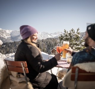 Ein Paar stoßt auf der winterlichen Terrasse der Alpe Dornach mit Blick auf schneebedeckte Alpen an – ein perfekter Moment auf der Alpe Dornach.