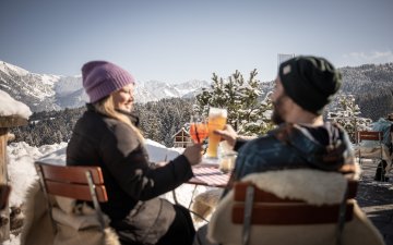 Ein Paar stoßt auf der winterlichen Terrasse der Alpe Dornach mit Blick auf schneebedeckte Alpen an – ein perfekter Moment auf der Alpe Dornach.