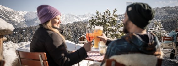 Ein Paar stoßt auf der winterlichen Terrasse der Alpe Dornach mit Blick auf schneebedeckte Alpen an – ein perfekter Moment auf der Alpe Dornach.