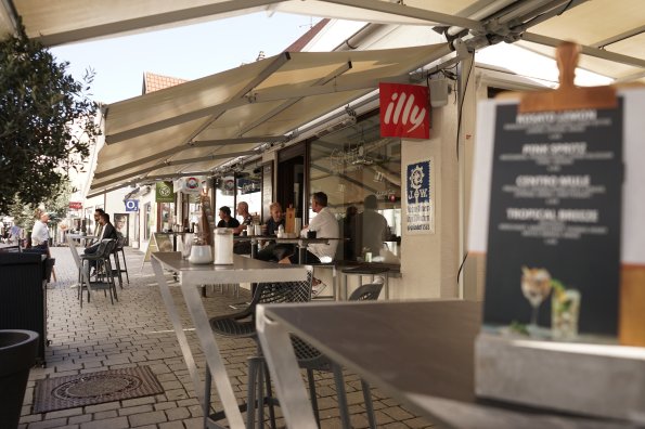 Gäste genießen entspannt Getränke im Außenbereich eines Cafés in einer lebhaften Stadtstraße an einem sonnigen Tag.