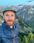 Ein Mann steht in den Bergen und lächelt. Er trägt eine Jacke und eine Kappe. Die Landschaft ist bergig und grün.