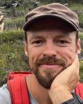 Mann macht ein Selfie mit einem Hund in den Bergen Ein Mann steht im Vordergrund und macht ein Selfie. Hinter ihm steht ein Hund auf einem Hügel in den Bergen.