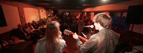Zwei Musiker stehen auf der Bühne und spielen vor vielen Zuschauern in einem kleinen Raum. Die Menschen hören aufmerksam zu.