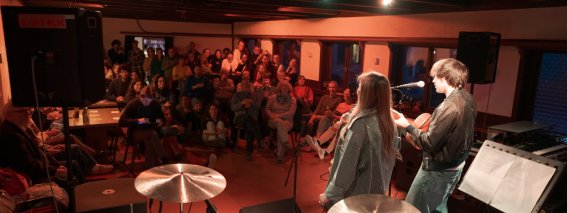 Ein Musiker und eine Musikerin spielen auf der Bühne vor einem Publikum in einem Raum. Die Zuschauer hören aufmerksam zu.
