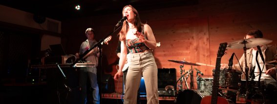 Eine Frau singt auf einer Bühne in einer Bar. Die Band spielt Instrumente und das Publikum hört zu.
