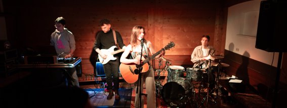 Eine Band spielt live auf der Bühne. Die Sängerin singt und spielt Gitarre. Es gibt einen Bassisten, einen Schlagzeuger und einen DJ.