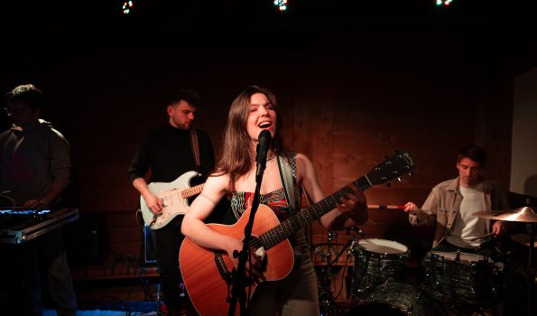 Eine Band tritt in einem kleinen Club auf. Die Sängerin spielt Gitarre und singt. Die Band hat mehrere Instrumente.