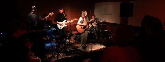 Eine Band spielt Live-Musik in einem kleinen Club. Die Atmosphäre ist lebendig und die Zuschauer genießen die Darbietung.