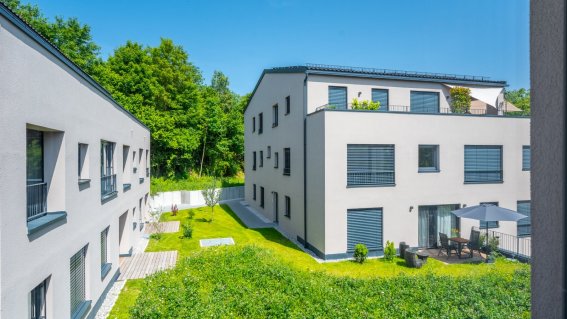 Zwei moderne Gebäude stehen in einer grünen Umgebung mit Gärten und Bäumen. Der Himmel ist klar und blau.