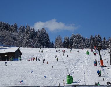 Auf einem schneebedeckten Hang tummeln sich viele Wintersportler beim Skifahren und Rodeln in der klaren Wintersonne.