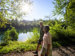 Zwei Personen gehen am Ufer eines Sees, umgeben von üppigem Grün, während die Sonne scheint.