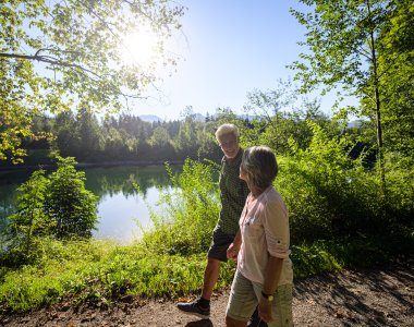 Zwei Personen gehen am Ufer eines Sees, umgeben von üppigem Grün, während die Sonne scheint.