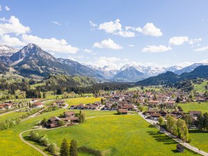 Grünes Tal mit weiten Wiesen und schneebedeckten Bergen, unter blauem Himmel, das Dorf erstrahlt im frischen Frühlingslicht.