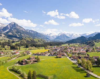 Grünes Tal mit weiten Wiesen und schneebedeckten Bergen, unter blauem Himmel, das Dorf erstrahlt im frischen Frühlingslicht.