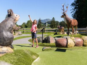 Ein Kind spielt Miniaturgolf in einer landschaftlich schönen Anlage mit Tierfiguren im Hintergrund an einem sonnigen Tag.