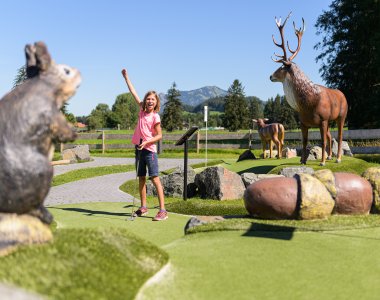 Ein Kind spielt Miniaturgolf in einer landschaftlich schönen Anlage mit Tierfiguren im Hintergrund an einem sonnigen Tag.