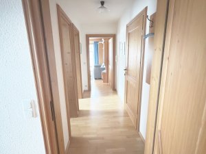 Der Flur zeigt Holzflächen und Türen. Im Hintergrund ist ein Raum mit Licht und Möbeln zu sehen.