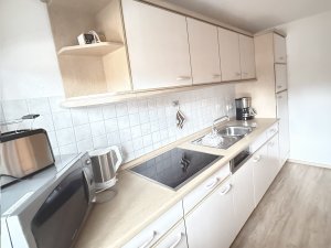 In der Küche stehen ein Herd, eine Spülmaschine und ein Kühlschrank. Die Wände sind hell, und der Boden ist aus Holz.