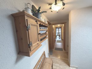 Der Flur zeigt ein Holzregal mit Geschirr und Pflanzen. Es gibt eine Tür am Ende des Flurs und eine Deckenlampe.