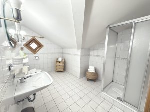 Das Badezimmer hat eine Dusche, ein Waschbecken und einen Aufbewahrungskorb. Es ist hell und hat weiße Fliesen.