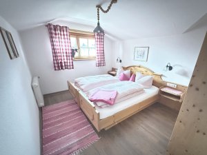 In einem hellen Zimmer stehen zwei Betten mit rosa Decken. Die Fenster haben rot-weiß karierte Vorhänge.