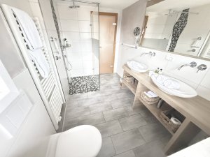 Das Badezimmer zeigt eine Dusche, zwei Waschbecken und einen Handtuchhalter. Alles ist hell und ordentlich eingerichtet.