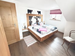 Das Zimmer zeigt ein Holzbett mit Kissen und eine Holzwand mit Kühen. Ein Stuhl steht neben dem Bett in einem hellen Raum.