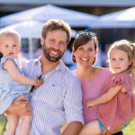 Eine Familie steht draußen und lächelt. Zwei Kinder sitzen in den Armen der Eltern. Es ist hell und sonnig.
