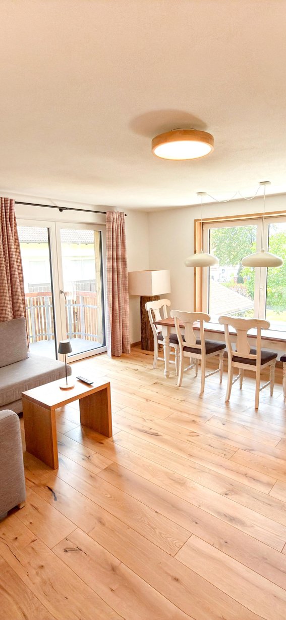 Das Wohnzimmer hat ein Sofa, einen Tisch und einen Esstisch. Es gibt große Fenster mit Blick ins Freie.