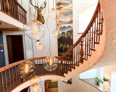 Das Treppenhaus zeigt eine Holztreppe und runde Lampen. An der Wand hängt ein Geweih. Fenster lassen Licht herein.