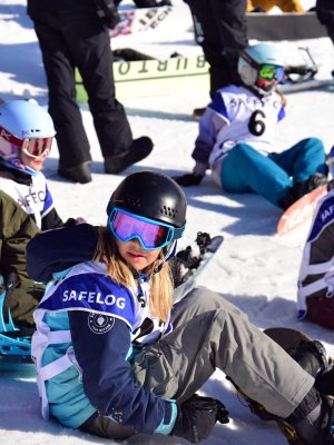 Eine Gruppe von Kindern sitzt auf dem Schnee und übt das Snowboarden in einem sonnigen Skigebiet.