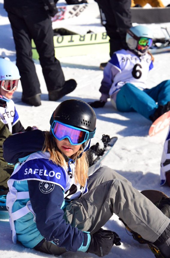 Eine Gruppe von Kindern sitzt auf dem Schnee und übt das Snowboarden in einem sonnigen Skigebiet.