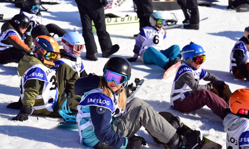 Eine Gruppe von Kindern sitzt auf dem Schnee und übt das Snowboarden in einem sonnigen Skigebiet.