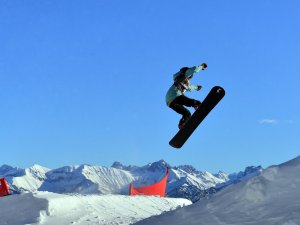 Ein Snowboardfahrer springt hoch über einen Schneehügel in einer winterlichen Berglandschaft. Es ist ein klarer, sonniger Tag.